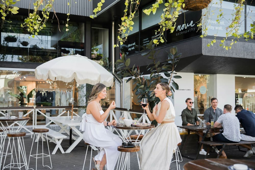 two women having wine and dinner outside a restaurant. group of men in background.