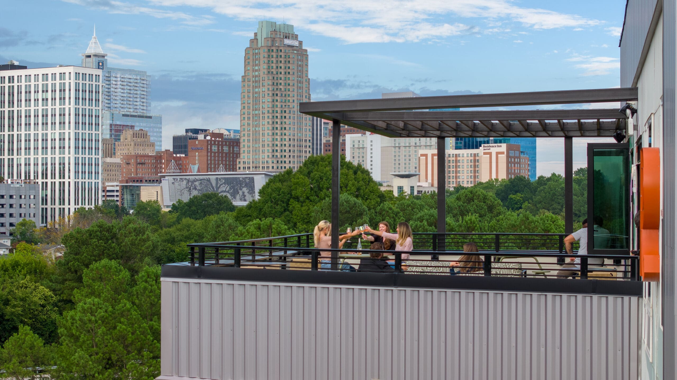 woman cheers win glasses on rooftop lounge. aerial view over downtown raleigh.